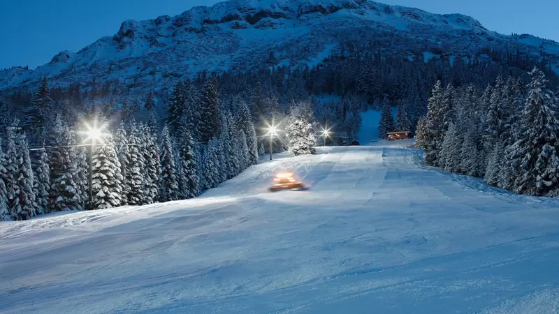 Blick über die beleuchtete Skipiste bei Nacht mit einer verschneiten Berglandschaft im Hintergrund.