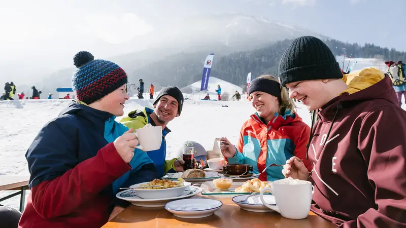 Eine Familie bei der Einkehr an der Skipiste, im Hintergrund eine verschneite Landschaft.
