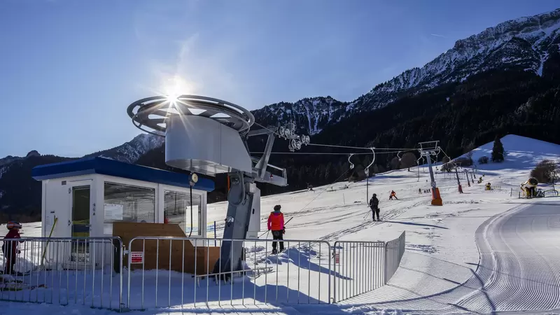 Skifahrer fahren mit einem Tellerlift, im Hintergrund ist eine weite Schneelandschaft zu sehen.